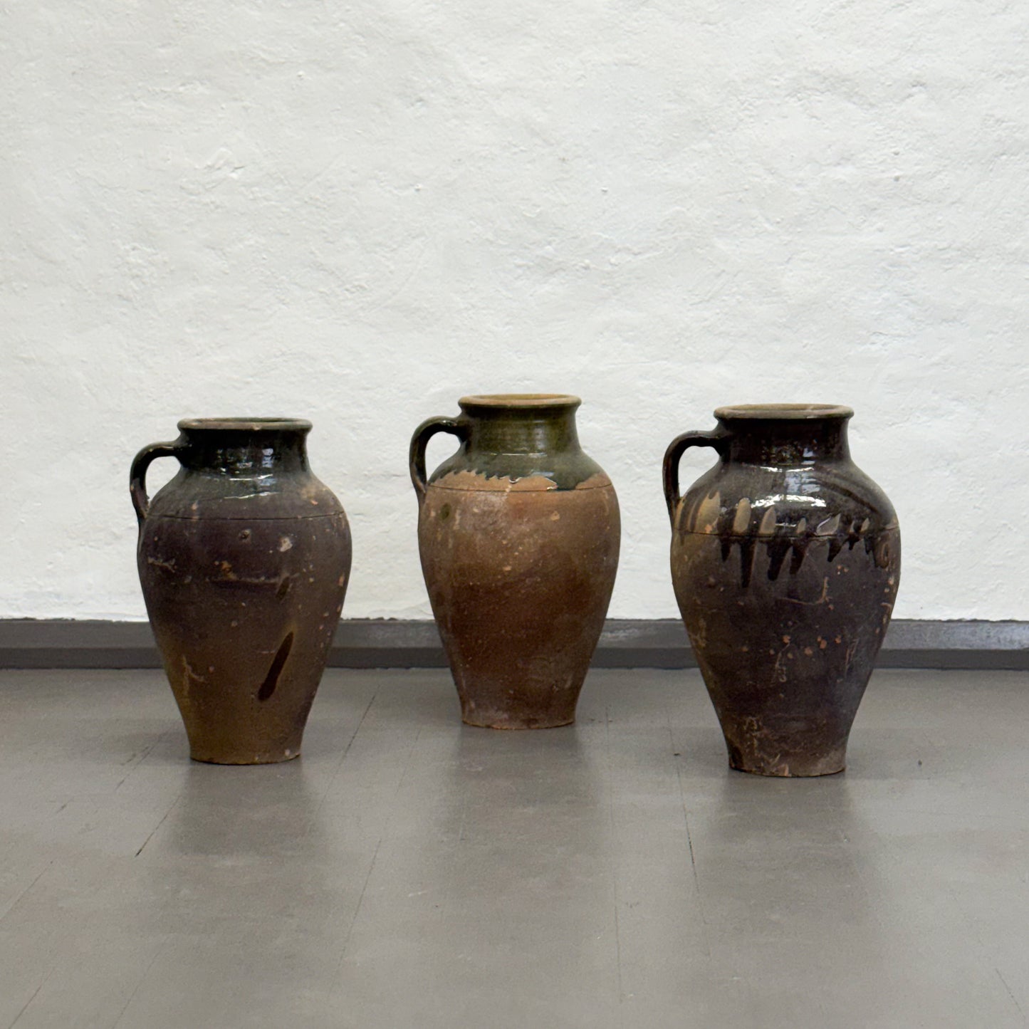 Three rustic clay pots with handles on a plain floor against a textured wall.