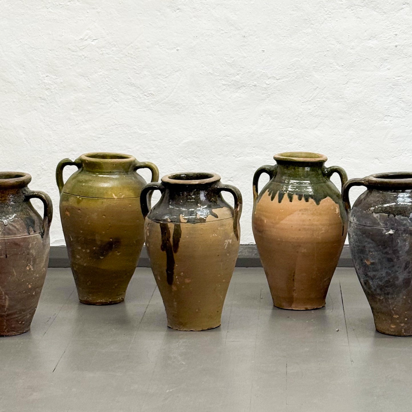 Set of five ceramic jars with different brown and green glaze finishes on a white brick wall background.
