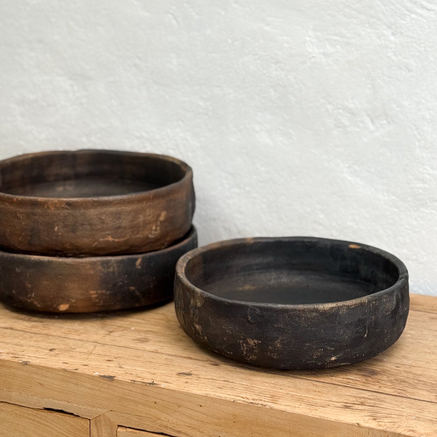 Three rustic wooden bowls on a wooden surface with a textured white wall background.